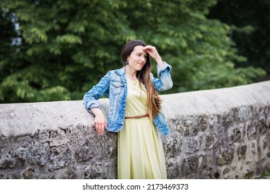 Lifestyle Portrait Of Young Stylish Woman With Long Brunette Hair Walking On The Street In Old Town, Wearing Yellow Dress And Denim Jacket