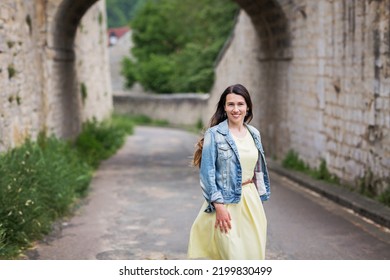 Lifestyle Portrait Of Young Stylish Woman With Long Brunette Hair Walking On The Street In Old Town, Wearing Yellow Dress And Denim Jacket