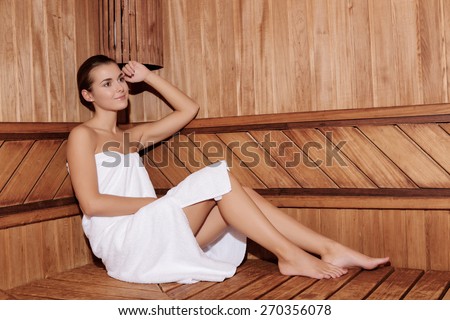 Lifestyle relaxation. Young dreamy woman in white towel sitting in a wooden sauna