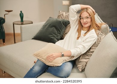 Lifestyle. Smiling middle aged mature woman looking at camera, happy lady in glasses posing at home indoor, positive female sitting at sofa in living room.