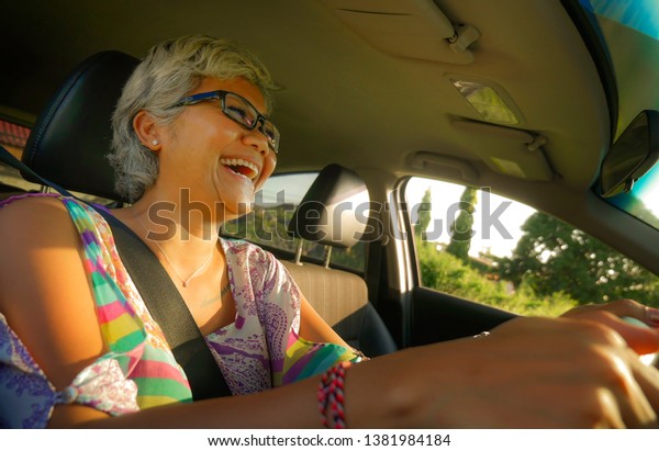 lifestyle Summer portrait of middle aged happy\
and attractive classy Asian Indonesian woman driving left hand car\
smiling cheerful and free on a sunny day in automobile rental and\
travel concept