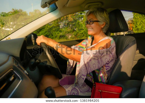 lifestyle Summer portrait of middle aged happy\
and attractive classy Asian Indonesian woman driving left hand car\
smiling cheerful and free on a sunny day in automobile rental and\
travel concept