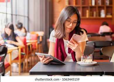  Lifestyle, Technology  Concept - Asian woman typing write message on smart phone and using tablet sitting at a table with cake in a modern cafe. 