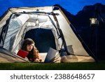 Lifestyle, Technology Concept - Woman holding book in her hands, working on laptop, lying in the camping tent and Camping lamp hanging on a steel stand. Landscape mountains at night. Rural scenery
