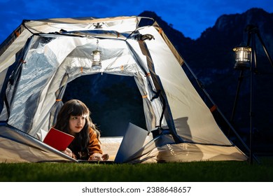 Lifestyle, Technology Concept - Woman holding book in her hands, working on laptop, lying in the camping tent and Camping lamp hanging on a steel stand. Landscape mountains at night. Rural scenery - Powered by Shutterstock