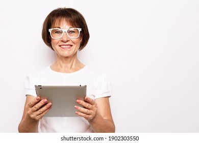 Lifestyle, Tehnology And Old People Concept: An Elderly Woman In A White T-shirt And Glasses Is Holding An IPad And Smiling. She Is Ready For New Knowledge.