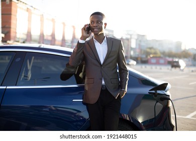 Lifestyle, Transportation And Ecology Concept. Handsome Young Black Man In Business Clothes, Talking Phone And Waiting For His Electric Car Recharging