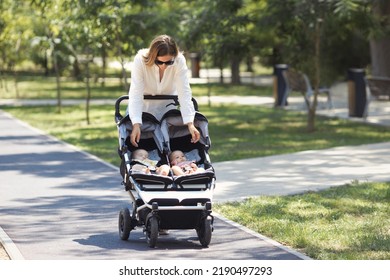 Lifestyle young casual caucasian mother with two little twin girls in a double baby stroller walks in city the park  - Powered by Shutterstock