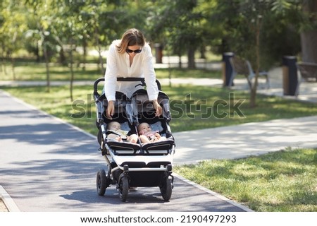 Lifestyle young casual caucasian mother with two little twin girls in a double baby stroller walks in city the park 