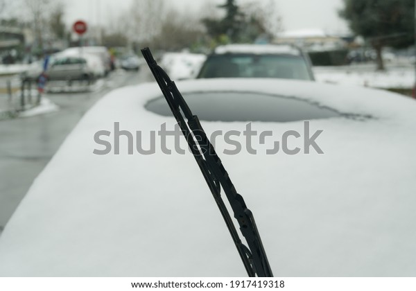 Lifted screen wiper before\
a car covered with snow. Day view of frozen windshield blade raised\
up against blurred background of a parked vehicle on a cold winter\
day.