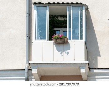 Light beige building features a small balcony. Double window has one pane open, casting a shadow. Wicker basket holds pink and purple flowers. Ideal for home decor or gardening themes