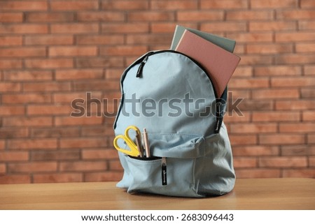 Light blue backpack with different stationery on wooden table against red brick wall