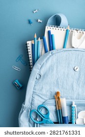 A light blue backpack stuffed with various school supplies such as pencils notebooks pens scissors paint brushes and paper clips set against a solid blue backdrop. This image is perfect for back to school themes and education.