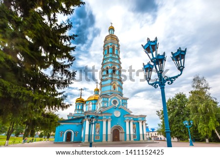 Light blue high church with golden domes surrounded by green trees under large rain clouds on the square. Cathedral of the Nativity of the Virgin, Ufa, Bashkortostan, Russia.