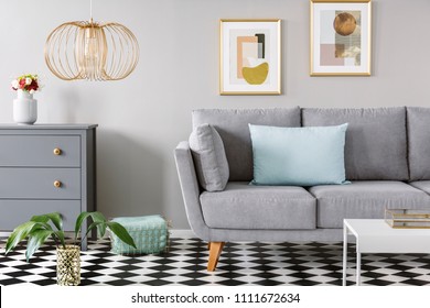 Light Blue Pillow Placed On Grey Couch In Bright Living Room Interior With Checkerboard Linoleum Floor, Fresh Flowers In Vase On Cupboard, Two Posters Hanging On The Wall And Gold Lamp