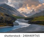 A light blue river winds through a dark gray mountain valley under a cloudy sky in Iceland. The image captures the stunning beauty of the Icelandic landscape.
