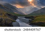 A light blue river winds through a valley in Iceland, surrounded by dark gray mountains and dramatic clouds with a bright orange glow breaking through.