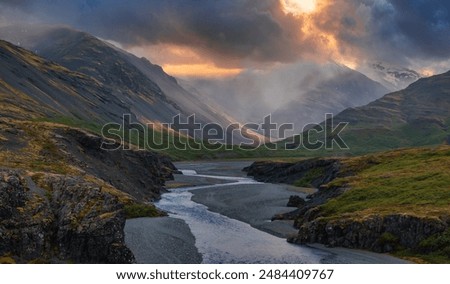 A light blue river winds through a valley in Iceland, surrounded by dark gray mountains and dramatic clouds with a bright orange glow breaking through.