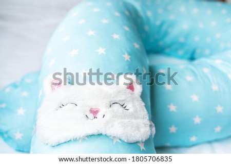 Light blue supporting pillow with stars on white sheet in bed for comfortable sleep of pregnant woman. Funny sleeping mask in form of bunny,cat with eyes,ears,mouth. Accessories for expectant mothers.