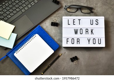 Light Box With The Text LET US WORK FOR YOU Near A Laptop And Notepad, Eyeglasses On A Gray Concrete Background. Flat Lay.