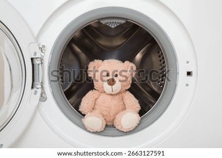 Light brown soft teddy bear sitting in white washing machine. Baby clothes or plush toys washing. Closeup. Front view.