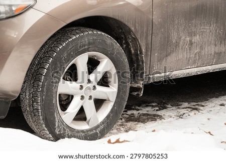 Light brown SUV front wheel and lower body covered in dirty snow and road salt, parked on a snowy road, depicting typical winter driving aftermath.