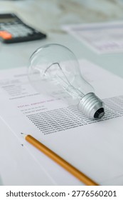 A light bulb lies on a stack of papers beside a pencil and a calculator on a desk during the day.