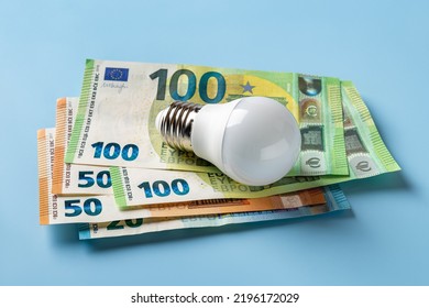 Light Bulb On 50 And 100 Euro Currency Banknotes Over Blue Background. High Energy Costs Concept. Power Price Rising In Europe. Soaring Of Electricity Bills. Energy Price Spike. Top View.