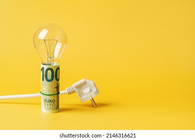 Light bulb on a rolled 100 euro banknotes and white power plug over yellow background. Copy space. Concept of increasing the electricity costs due to power crisis and inflation. Rising energy costs.