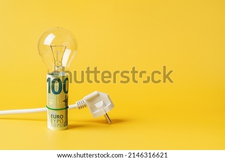Light bulb on a rolled 100 euro banknotes and white power plug over yellow background. Copy space. Concept of increasing the electricity costs due to power crisis and inflation. Rising energy costs.