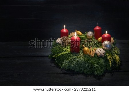 Light in the dark on third advent, natural green wreath with red candles, three are burning, Christmas decoration and cookies, dark wooden background, copy space, selected focus