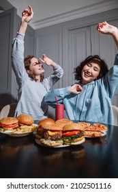 A light faded, two girls in casual clothes sitting at the black table dancing their hands up and eyes closed, pizza and burgers on the table