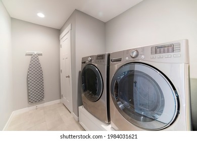 Light gray laundry room with tiles, washer, dryer and storage room. There is a white door beside the laundry machines on top of a white board, and a patterned ironing board hanging on the wall.