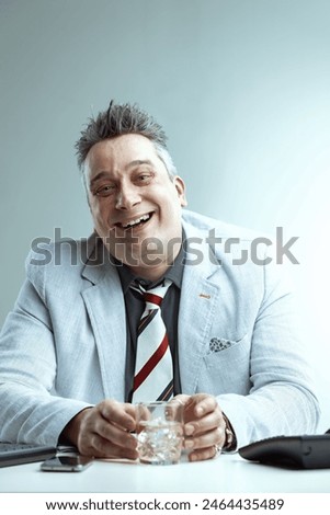 Light gray-haired man in a light gray blazer, dark gray shirt, and striped tie, holds a glass of alcohol with a smug expression, thinking it's normal at work, office setting