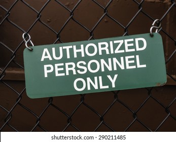 Стоковая фотография: Light green outdoor sign, slightly crooked, on black chain-link fence by dark red steel wall: AUTHORIZED PERSONNEL ONLY