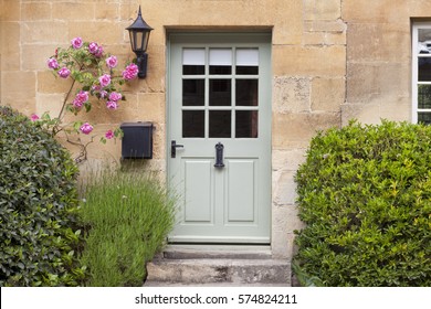 Light green wooden doors in an old traditional English lime stone cottage surrounded by climbing pink roses, lavender, on summer day