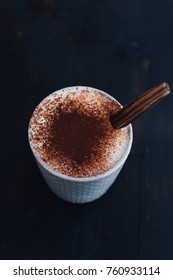 Light grey stoneware mug with stitched pattern filled with hot chocolate with slightly whipped cream and dusted with cocoa powder on black background and wooden spoon