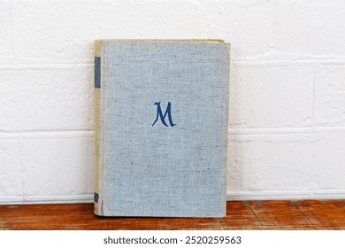 A light grey vintage book with a minimalist design, featuring a large, embroidered letter "M" on the front cover. The book is set against a white textured wall on a wooden surface. - Powered by Shutterstock