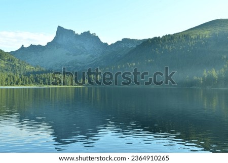 A light haze over the unsteady surface of a large lake in a dense coniferous forest at the foot of high peaked mountains. Lake Svetloe, Ergaki Nature Park, Krasnoyarsk Territory, Siberia, Russia.