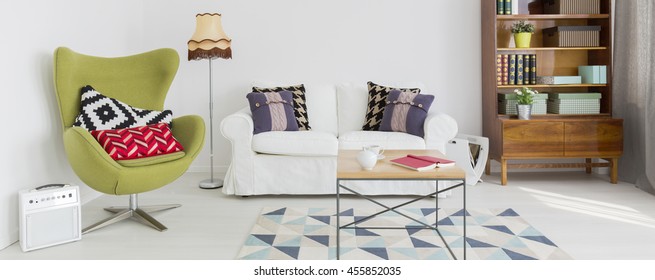 Light home interior with green egg chair, sofa, small table, wooden bookshelf and pattern carpet