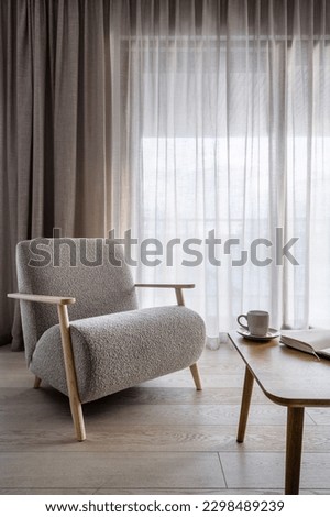 Light living space in modern apartment. Sophistication soft grey armchair against windows with linen curtains. Contemporary furniture and decor in comfortable room. Open book or diary on wooden table