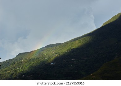 Light multicolored rainbow appearing above rural buildings among forest on hillside under overcast cloudy sky