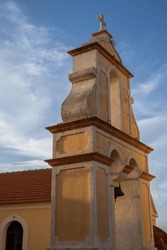 Light Orange Color Of The Facade Of Orthodox Church. Tradional Separate Belfry With A Cross. Blue Sky With White Clouds. Late Afternoon Sunshine. Acharavi, Corfu, Greece.