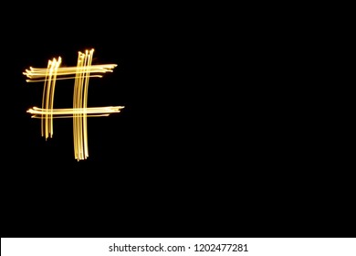 Light Painting Photography, Long Exposure, Gold Metallic Yellow Light Movement Depicting The Hashtag Symbol Against A Black Background.