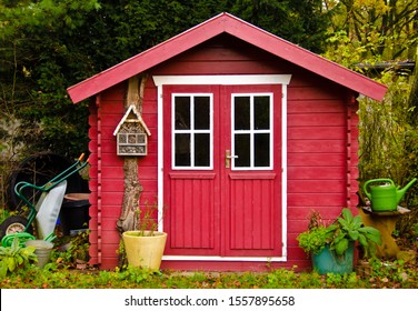 A light red small shet, gardenhouse, with some garden tools around it