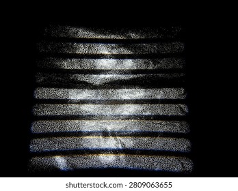 Light shines through a surface with grooves, casting lines and shapes in the darkness. The scene shows contrasts between light and shadow. This takes place at night.