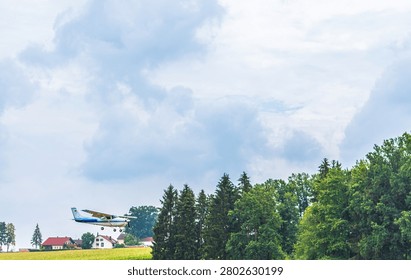 A light single engine airplane flies at low altitude over a rural landscape, approaching a small airfield. The scene shows green trees, farm fields and village houses under a cloudy summer sky with wide negative space.
