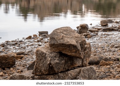 light sky and trees reflected in still water.  Still water reflecting trees, sky and large rocks in foreground.  Rocky shore foreground with greenery.   - Powered by Shutterstock