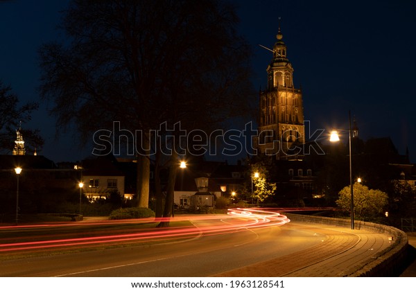 Light streaks of\
passing cars on meandering town road lit up by city lights with\
Walburgiskerk cathedral towering behind into the night. Long\
exposure blue hour\
cityscape.