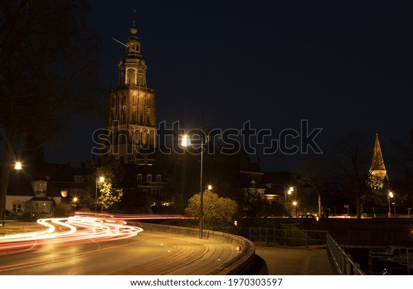 Light streaks of passing cars on meandering town\
road lit up by city lights with Walburgiskerk cathedral and\
Drogenapstoren towering\
behind